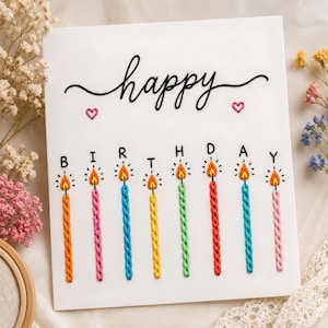 May include: A white greeting card featuring black embroidered text that reads happy, two small pink embroidered hearts, and the word BIRTHDAY in black letters. Below each letter is a colorful embroidered candle with a yellow and orange flame.