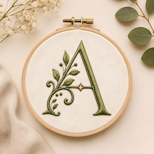 May include: An embroidery hoop containing a piece of off-white fabric with a large, dark green embroidered letter A. The letter is decorated with intricate green leaves, swirling vines, and a small gold star detail at the center crossbar.