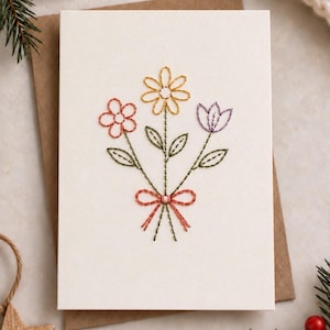 Op de afbeelding: Een rechthoekige witte wenskaart met een handgeborduurd ontwerp van drie bloemen in geel, koraal en paars met groene stelen en bladeren, onderaan samengebonden met een koraalkleurige geborduurde strik.