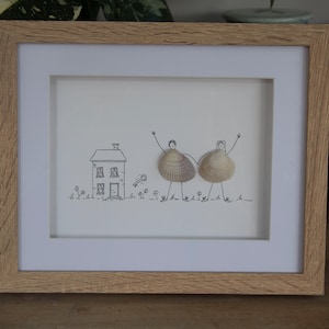 Puede incluir: Una obra de arte enmarcada que presenta un dibujo lineal de una casa y dos figuras con cuerpos de conchas marinas. Las figuras se toman de la mano junto a una ilustración de una llave. El marco es de madera clara con un paspartú blanco.