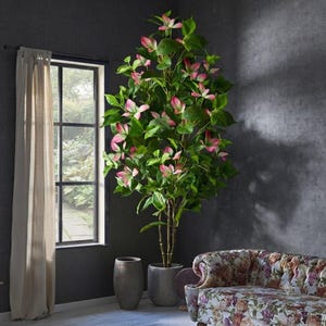 May include: A tall artificial dogwood tree with vibrant green leaves and delicate pink-tipped blossoms. The plant features multiple slender brown trunks and is housed in a simple, cylindrical gray planter, standing upright in a room.