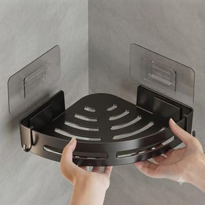 Puede incluir: Una estantería de esquina para ducha en color negro mate con borde frontal curvo y base ranurada para el drenaje. La estantería se fija a la pared mediante dos soportes adhesivos rectangulares transparentes con un mecanismo de enganche central.