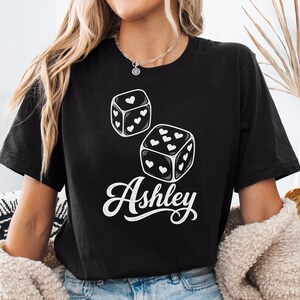 May include: A black short-sleeved t-shirt featuring a white graphic of two dice with heart-shaped pips instead of dots. Below the dice, the name Ashley is printed in a white cursive script font. The shirt has a crew neckline and a standard fit.