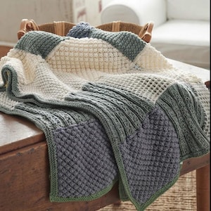 May include: A chunky knit blanket featuring a patchwork design of cream, sage green, and dark grey squares. The blanket displays various textures including waffle stitch, ribbed patterns, and bobble stitches, finished with a solid green border.