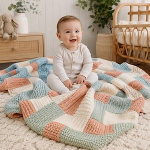 May include: A knitted baby blanket featuring a patchwork design of square blocks in soft shades of dusty rose, sage green, and cream. The blanket has a textured garter stitch pattern and is displayed spread out on a textured white rug.