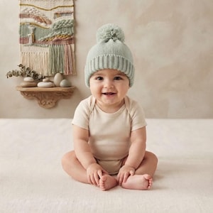 Pode incluir: Um gorro de malha verde-sálvia claro com um pompom a condizer no topo, apresentando uma bainha canelada e uma textura macia. O gorro é usado por um bebé que também veste um body liso de manga curta em bege claro, feito de tecido suave.