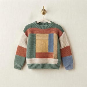 May include: A long-sleeve knit sweater featuring a color-blocked pattern of sage green, terracotta, mustard yellow, cream, and slate blue rectangular sections. The garment has a crew neckline, ribbed cuffs, and a ribbed hem, hanging on a gold hanger.