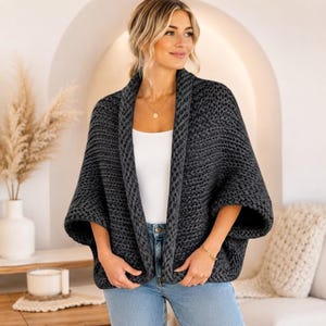 May include: A dark charcoal gray chunky knit cardigan with an open front, wide sleeves, and a thick ribbed collar. The garment is worn over a plain white top and light blue denim jeans, featuring a textured, heavy yarn construction throughout.