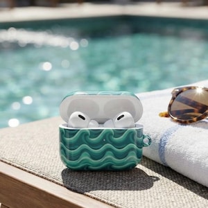 May include: A white wireless earbud charging case featuring a green and teal wavy pattern. The case is open, revealing two white earbuds inside. The case includes a small circular loop on the right side for attaching a lanyard or accessory.