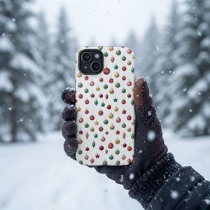 Può includere: Una cover per smartphone bianca con un motivo ripetuto di piccoli ornamenti natalizi colorati in rosso, verde e oro, tra cui palline rotonde, stelle e forme a goccia, con un ritaglio nero per l'obiettivo della fotocamera.