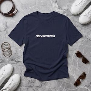 Pode incluir: Uma t-shirt azul-marinho de manga curta com a palavra NEVERMIND impressa em branco no centro do peito. A peça tem gola redonda e corte padrão, apresentada na horizontal com um cinto castanho, ténis brancos e óculos de sol.