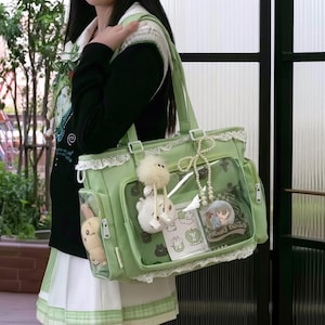 May include: A light green tote bag featuring a clear front pocket, white lace trim along the edges, and two shoulder straps. The bag is decorated with a fluffy white charm, a pearl bead string, and various small character-themed stickers and cards.