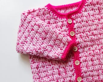 Puede incluir: Un cárdigan de bebé de punto de crochet rosa y blanco con botones de madera.