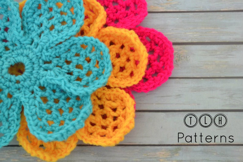 May include: A multi-colored crocheted flower with teal, yellow, and pink petals. The flower is on a wooden surface with the text "TLH Patterns" below it.