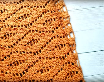 Puede incluir: Una manta de ganchillo de color naranja cálido con un patrón de encaje. La manta está doblada y colocada sobre una superficie de madera blanca.