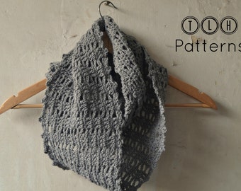 Puede incluir: Un pañuelo infinito gris tejido a ganchillo cuelga de una percha de madera. El pañuelo tiene un diseño abierto y calado y está hecho de un hilo suave y texturizado. El texto "TLH Patterns" es visible en la esquina superior derecha.