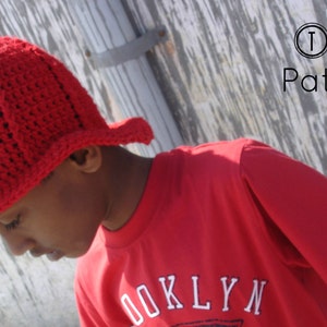 May include: A young person wearing a red crocheted firefighter hat with a white and black emblem. The hat has a wide brim and is pulled down over the person's forehead. The person is wearing a red t-shirt with the word "BROOKLYN" printed on the front.