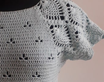 Puede incluir: Una blusa de crochet azul claro con un delicado patrón de encaje en las mangas y un escote redondo simple.