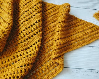 Puede incluir: Un chal de ganchillo amarillo mostaza con un borla de flecos. El chal está hecho con un delicado patrón de punto abierto.