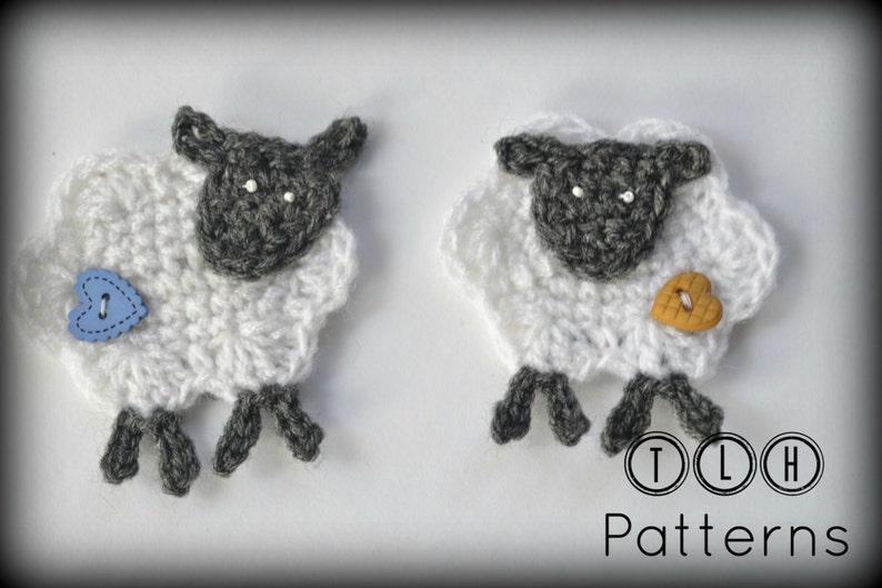 Op de afbeelding: Twee gehaakte schapen met witte wol en grijze gezichten en poten. E&eacute;n schaap heeft een blauwe hartvormige knoop, de andere een bruin hartvormige knoop. De tekst "TLH Patterns" staat in de rechter benedenhoek.