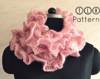 May include: A pink crocheted ruffle scarf on a black mannequin. The scarf is made with a soft, fluffy yarn and has a delicate, feminine look. The text "TLH Patterns" is visible in the top right corner of the image.