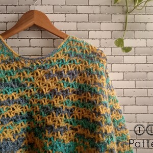 May include: A crocheted poncho with a striped pattern in shades of yellow, green, and blue. The poncho is hanging on a wooden hanger against a white brick wall.