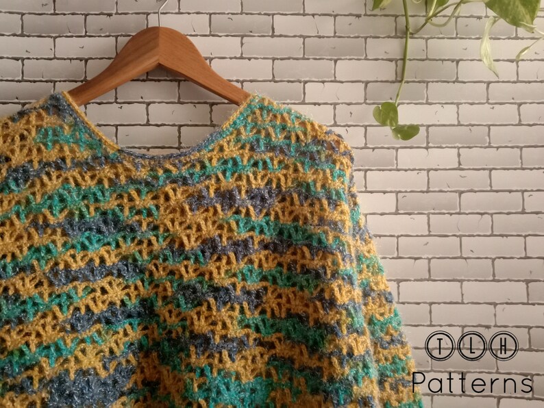 May include: A crocheted poncho with a striped pattern in shades of yellow, green, and blue. The poncho is hanging on a wooden hanger against a white brick wall.