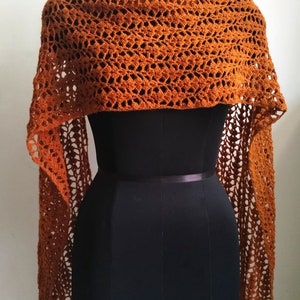 May include: A burnt orange crochet shawl on a black mannequin. The shawl features an open, lacy pattern and long, fringed edges. It is draped over the mannequin's shoulders, highlighting its texture and shape. The shawl is a warm, earthy tone.
