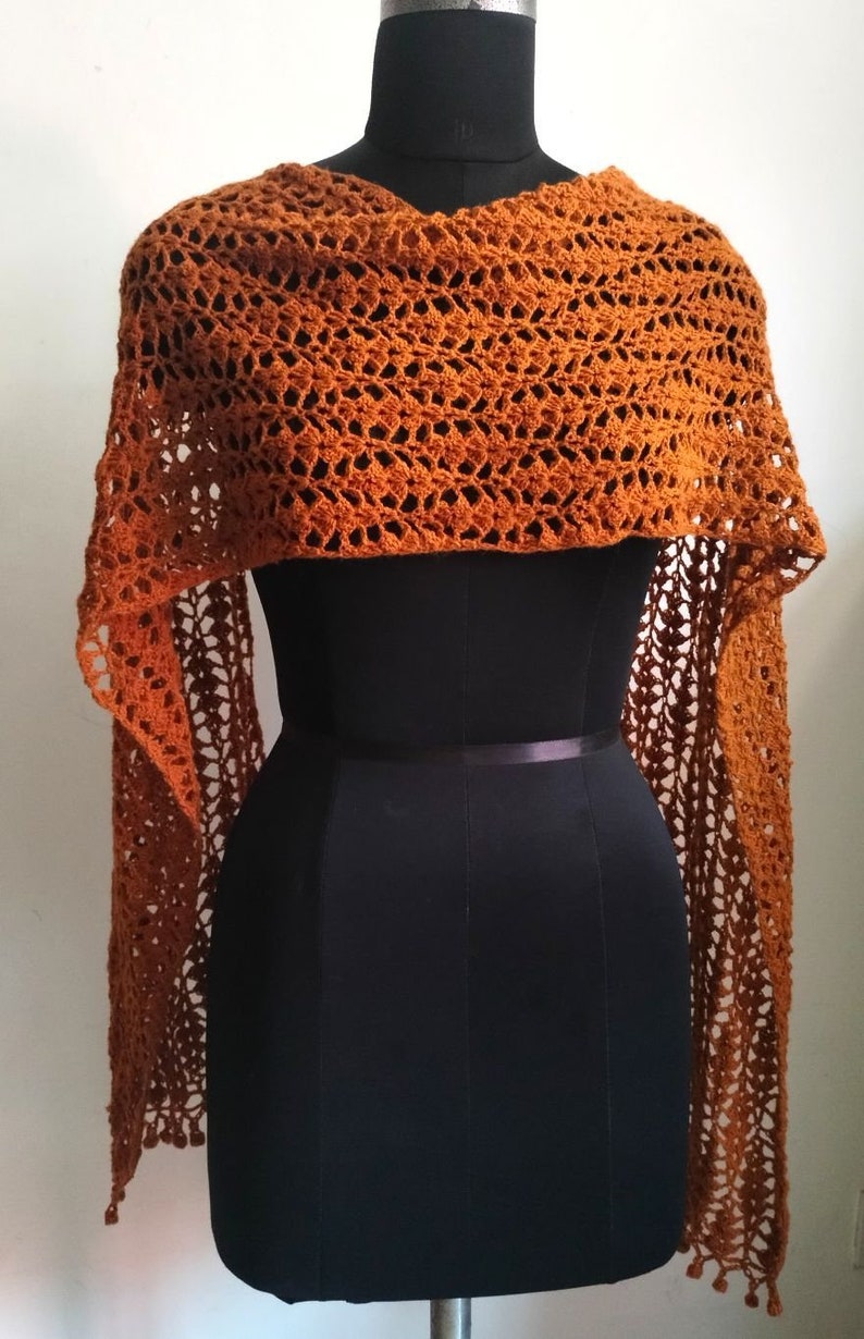 May include: A burnt orange crochet shawl on a black mannequin. The shawl features an open, lacy pattern and long, fringed edges. It is draped over the mannequin's shoulders, highlighting its texture and shape. The shawl is a warm, earthy tone.