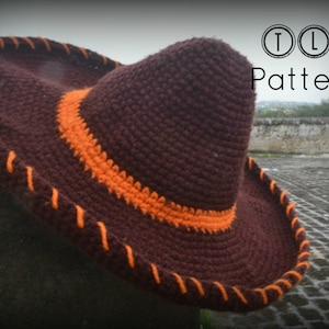 May include: A brown crocheted hat with an orange trim. The hat has a wide brim and a tall crown. The hat is sitting on a cobblestone surface.