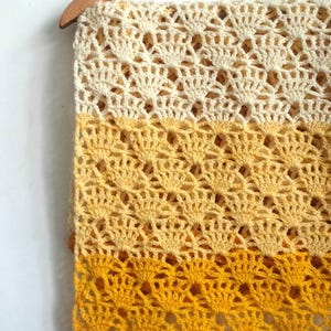 Peut inclure: Écharpe en crochet faite à la main, à tissage ouvert, dans un dégradé de jaunes, allant de la crème au jaune doré, jusqu'à un orange profond. L'écharpe est présentée sur un cintre en bois sur fond blanc.