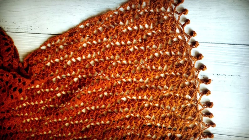 May include: A handcrafted, open-weave crochet shawl in shades of orange and brown. The shawl has a delicate, lacy pattern with a scalloped edge. The texture and colour suggest a warm, cosy accessory.