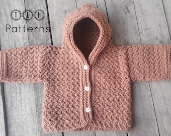 Könnte beinhalten: Eine braune gehäkelte Kapuzenjacke mit weißen Knöpfen. Die Jacke ist mit einem strukturierten Stichmuster gearbeitet.