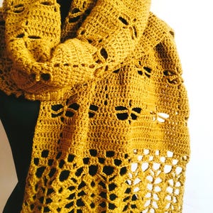 Puede incluir: Bufanda de ganchillo amarillo mostaza con patrones calados de libélulas y hojas. La bufanda está drapeada sobre una forma negra, mostrando los detalles del diseño hecho a mano. La textura y el color crean una estética cálida.