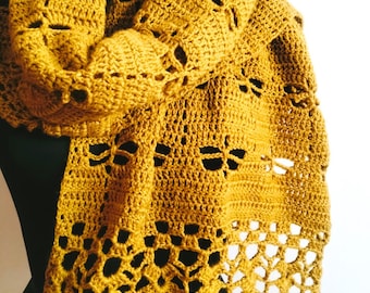 Puede incluir: Bufanda de ganchillo amarillo mostaza con patrones calados de libélulas y hojas. La bufanda está drapeada sobre una forma negra, mostrando los detalles del diseño hecho a mano. La textura y el color crean una estética cálida.