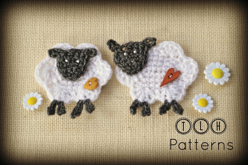 Op de afbeelding: Twee handgemaakte gehaakte schapen met witte wol en grijze gezichten, poten en oren. E&eacute;n schaap heeft een houten knoop, de andere een houten hart. Twee madeliefjesknopen en de tekst "TLH Patterns" zijn ook zichtbaar.