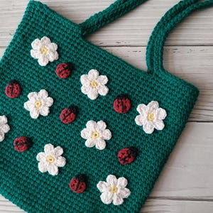 Könnte beinhalten: Eine handgefertigte, smaragdgrüne Häkeltasche mit weißen Blumen und roten Marienkäfer-Applikationen. Die Tasche hat zwei passende Griffe und ein strukturiertes Stichmuster, ideal für den Transport von Gegenständen.