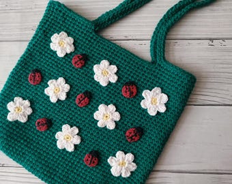 May include: A handmade, emerald green crochet tote bag adorned with white flowers and red ladybug appliqués. The bag features two matching handles and a textured stitch pattern, perfect for carrying essentials.
