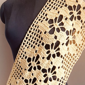 Puede incluir: Un chal de ganchillo crema de tejido abierto drapeado sobre un maniquí negro. El chal presenta un patrón floral con espacios abiertos, creando un diseño delicado y aireado. La textura es visible, destacando la calidad artesanal de la prenda.