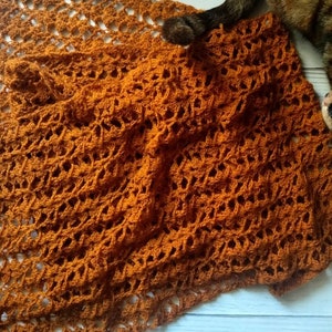 May include: An orange crocheted shawl with an openwork design. The shawl has a repeating pattern and is finished with tassels along the edges. The warm colour and intricate details make it a stylish accessory. The shawl is made of soft yarn.