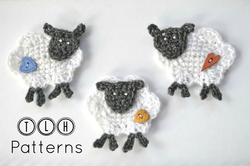 Op de afbeelding: Drie handgemaakte gehaakte schapen met witte wol en grijze gezichten en poten. Elk schaap heeft een knoop in de vorm van een hart in blauw, oranje of bruin. De tekst "TLH Patterns" staat onderaan.