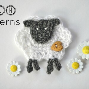 Op de afbeelding: Een gehaakt schaap in wit en grijs, met een hartvormige knoop en drie madeliefjesversieringen. De afbeelding bevat de tekst "TLH Patterns" in een cirkelvormig ontwerp.