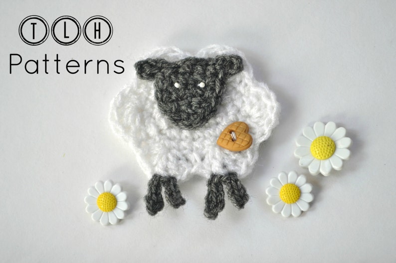 Op de afbeelding: Een gehaakt schaap in wit en grijs, met een hartvormige knoop en drie madeliefjesversieringen. De afbeelding bevat de tekst "TLH Patterns" in een cirkelvormig ontwerp.
