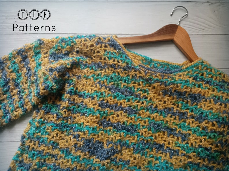 May include: A crocheted sweater in a striped pattern of mustard yellow, teal, and gray yarn. The sweater is hanging on a wooden hanger.
