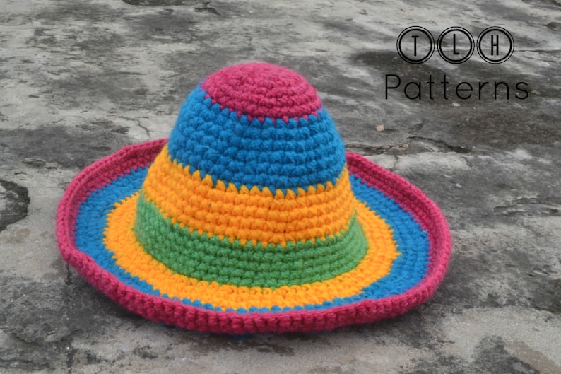 Pode incluir: Um chap&eacute;u de sol de croch&ecirc; colorido com listras rosa, azul, amarelo e verde. O chap&eacute;u est&aacute; sobre uma superf&iacute;cie de concreto cinza.