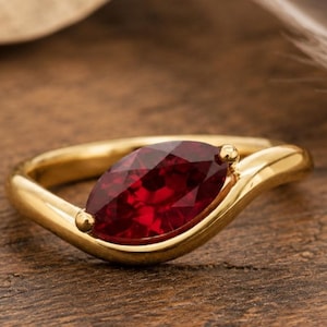 May include: A gold ring featuring a single marquise-cut deep red gemstone set in a smooth, curved band. The metal has a polished finish, and the stone is positioned centrally within the setting, showcasing a faceted surface and rich color.
