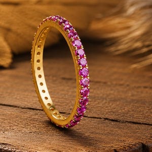 Könnte beinhalten: Ein dünner Goldring mit einer durchgehenden Reihe kleiner, runder, leuchtend pinker Edelsteine am äußeren Rand. Die Innenseite des Goldbandes weist eine Reihe kleiner, kreisförmiger Aussparungen entlang des gesamten Umfangs des Rings auf.