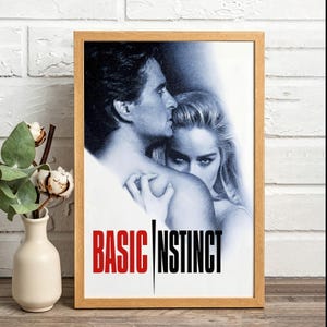 May include: A framed movie poster for Basic Instinct featuring a black and white photographic image of a man and a woman in close proximity. The title Basic Instinct is printed in bold red and black capital letters at the bottom of the poster.