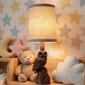May include: A table lamp featuring a base sculpted as a brown owl perched on a rock, topped with a cylindrical lampshade made of light brown textured fabric with dark gray trim at the top and bottom edges, emitting a warm, soft glow.