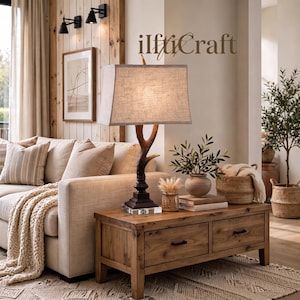 May include: A table lamp with a dark brown antler-shaped base, a rectangular linen lampshade, and a clear glass square pedestal. The lamp features the text ilftiCraft printed above the shade. It sits on a wooden coffee table with two drawers.
